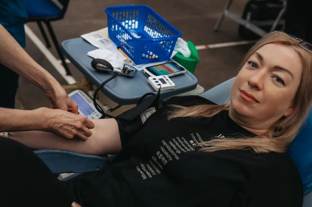 A woman lies back in a donation chair with her arm extended while a clinician prepares it, beside a table of blood pressure and testing equipment