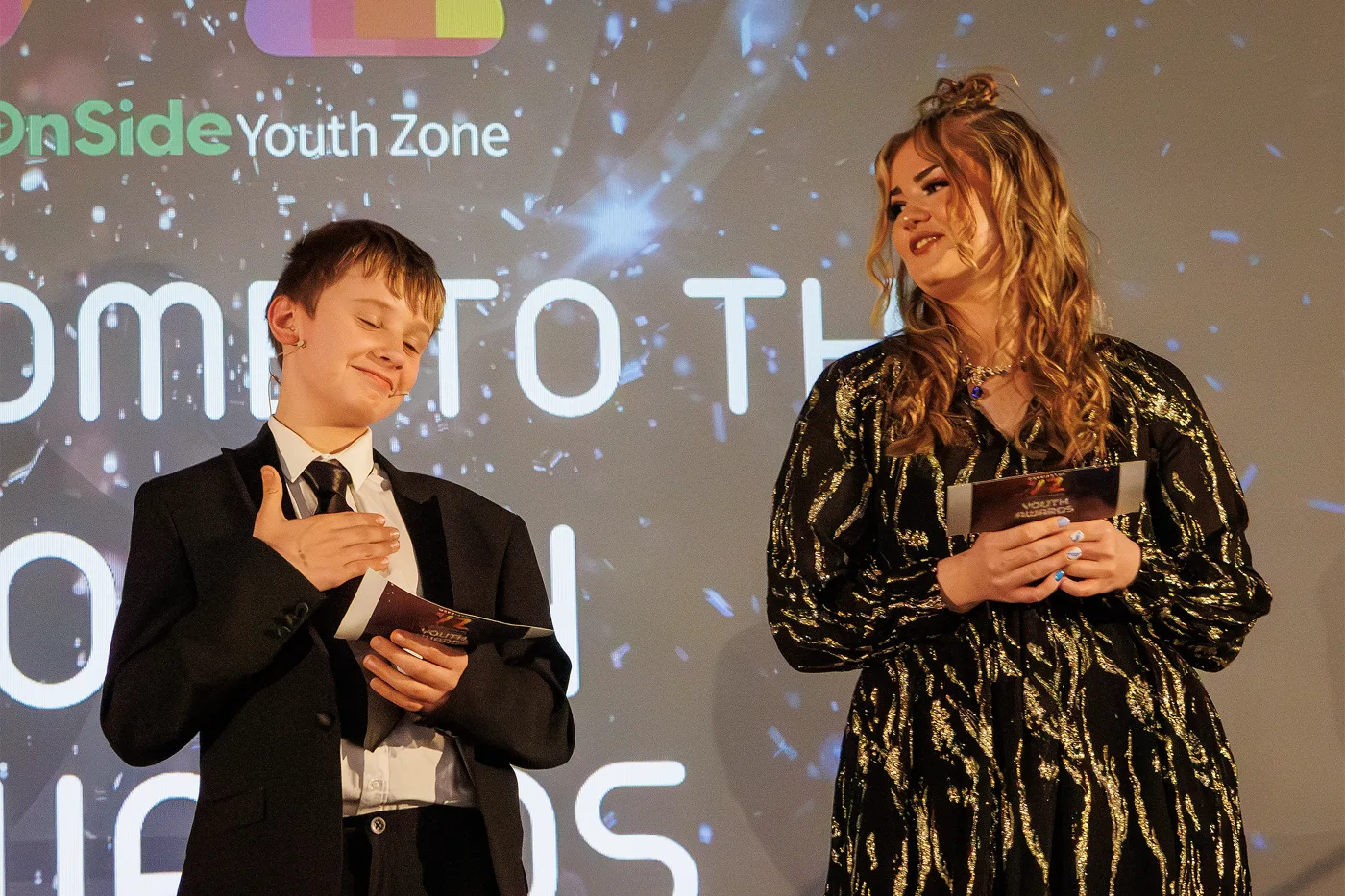 A boy in a suit with his hand on his chest and a young woman in a black and gold dress holding cue cards on stage, in front of a screen showing the OnSide Youth Zone logo